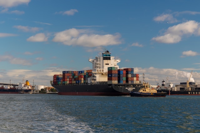 A large container ship, fully loaded with colorful shipping containers, being guided into a busy port by a smaller tugboat under a blue sky with clouds.