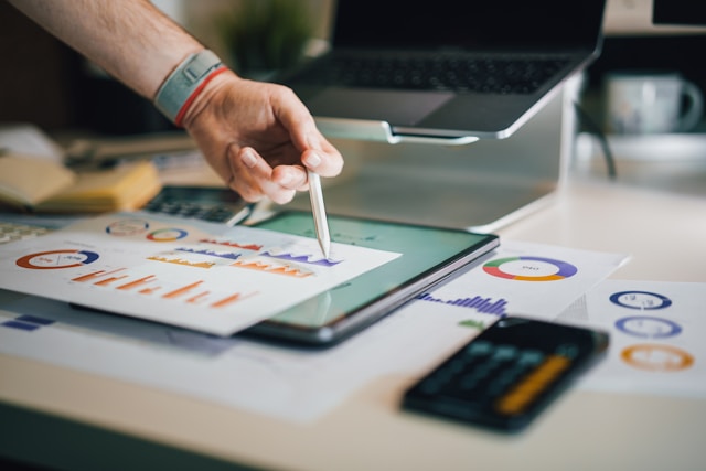 A person's hand pointing with a stylus at charts and graphs displayed on a tablet and paper, surrounded by a laptop and calculator, symbolizing data analysis, strategic planning, and logistics optimization.