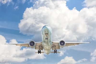 A large commercial passenger jet viewed from directly below, with its landing gear down, against a bright blue sky with white clouds, symbolizing global air freight, international connectivity, and worldwide reach.